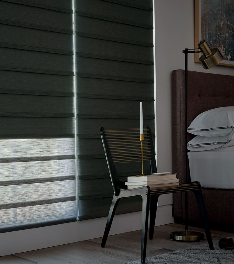 bedroom darkened by roller shades with woven backed chair with stacks of books