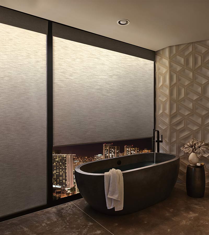 a modern bathroom with a tub next to large windows covered by illuminated privacy shades with warm light